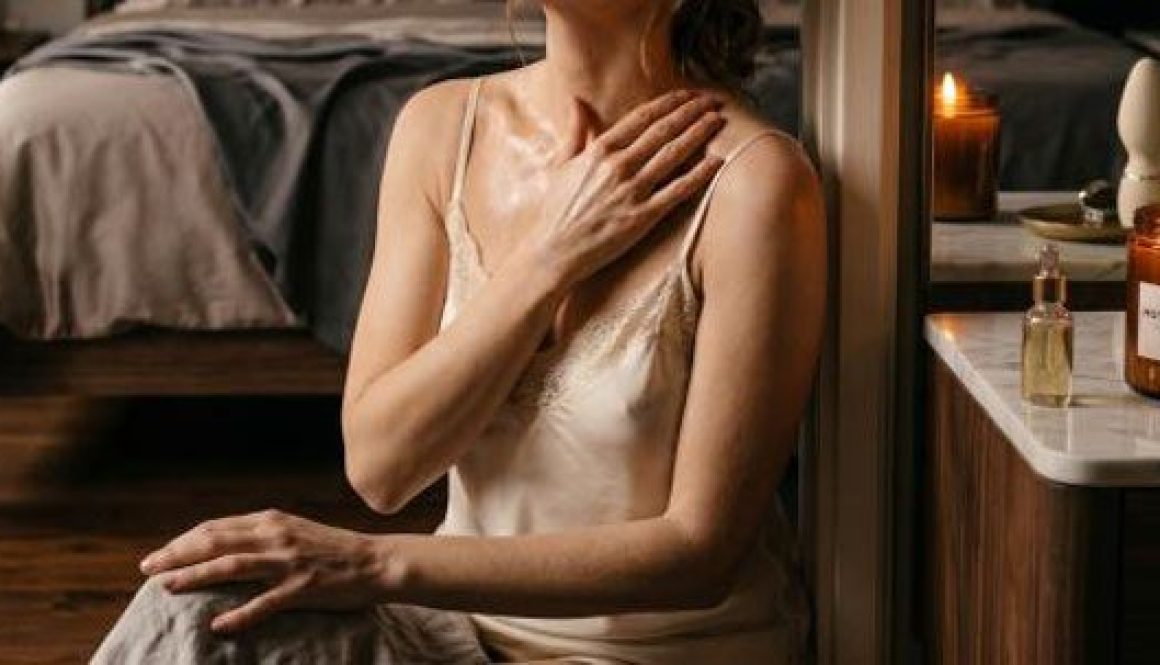 Donna in abito di seta che applica olio sul décolleté accanto a candela profumata al cedro e muschio bianco su marmo, rituale sensoriale consapevole —Eros Atelier